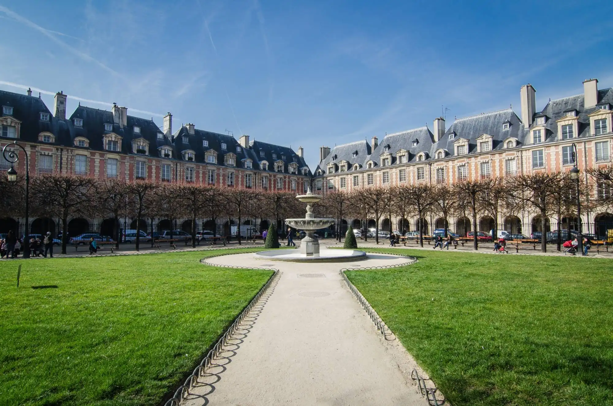 Place des Vosges dans le Marais : jardin, fontaine et pavillons de brique et de pierre sous un ciel bleu