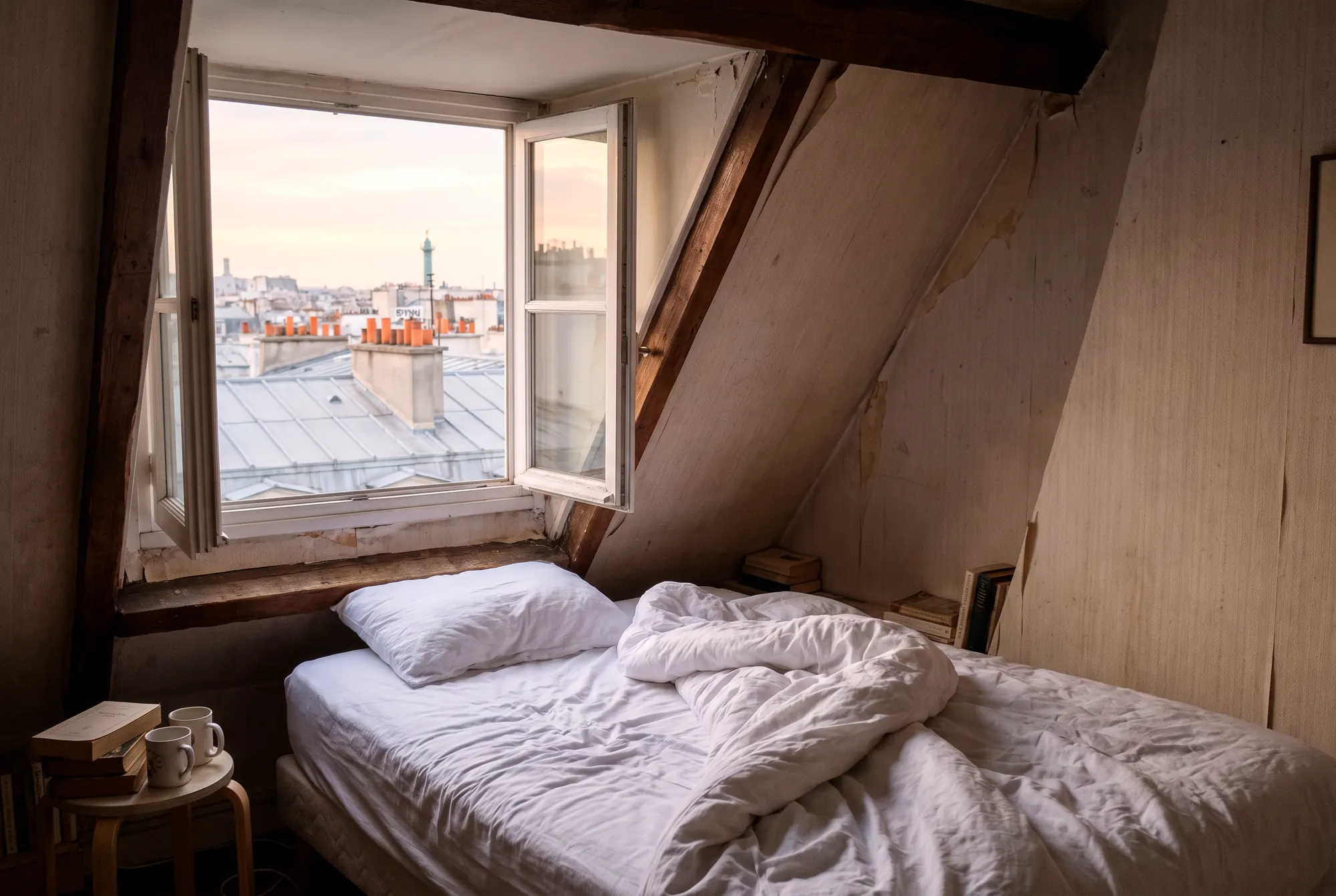 Chambre mansardée d’un appartement parisien, fenêtre ouverte sur les toits en zinc, lit défait après le départ des voyageurs
